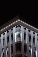 A modern building facade with a geometric design. The structure features a series of vertical panels and windows illuminated by subtle lighting, set against a dark night sky. The clean lines and lighting create a sleek, contemporary look.