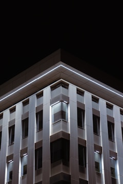 A modern building facade with a geometric design. The structure features a series of vertical panels and windows illuminated by subtle lighting, set against a dark night sky. The clean lines and lighting create a sleek, contemporary look.
