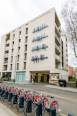 A modern multi-story building features unique vertical blue and teal art installations on its facade. A row of red rental bicycles lined up in docking stations is visible in the foreground, with trees and other buildings partially seen in the background.