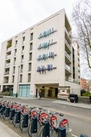 A modern multi-story building features unique vertical blue and teal art installations on its facade. A row of red rental bicycles lined up in docking stations is visible in the foreground, with trees and other buildings partially seen in the background.