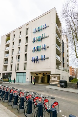 A modern multi-story building features unique vertical blue and teal art installations on its facade. A row of red rental bicycles lined up in docking stations is visible in the foreground, with trees and other buildings partially seen in the background.