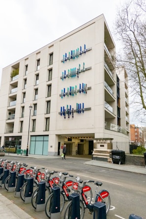A modern multi-story building features unique vertical blue and teal art installations on its facade. A row of red rental bicycles lined up in docking stations is visible in the foreground, with trees and other buildings partially seen in the background.