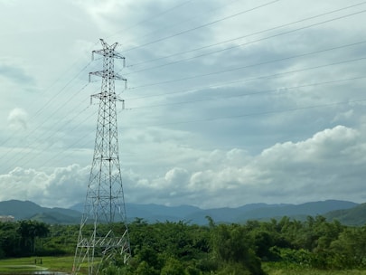 A tall metal transmission tower stands amidst a lush green landscape. The tower extends upward into a cloudy sky, with power lines stretching horizontally across the image. In the background, a range of hills creates a natural backdrop, enhancing the contrast with the industrial structure.