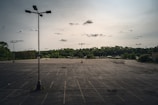 An expansive, empty parking lot with several tall light poles scattered throughout the space. The background features a dense tree line under a cloudy sky. Faded parking lines and dark cracks are visible on the asphalt surface.