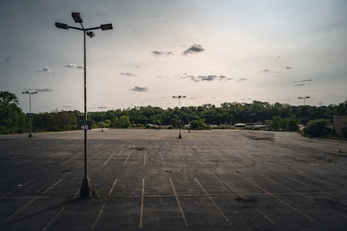 An expansive, empty parking lot with several tall light poles scattered throughout the space. The background features a dense tree line under a cloudy sky. Faded parking lines and dark cracks are visible on the asphalt surface.