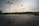 An expansive, empty parking lot with several tall light poles scattered throughout the space. The background features a dense tree line under a cloudy sky. Faded parking lines and dark cracks are visible on the asphalt surface.