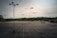 An expansive, empty parking lot with several tall light poles scattered throughout the space. The background features a dense tree line under a cloudy sky. Faded parking lines and dark cracks are visible on the asphalt surface.