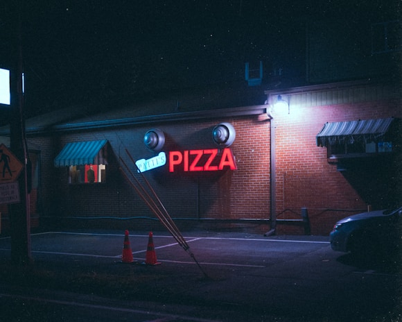 A nighttime scene of a brick building with a neon red sign that reads 'PIZZA' and a smaller sign above it. There are lights casting blue hues on the building. Two conical traffic cones are placed next to a slanted post in front of the building. There is also a car partially visible on the right side and a person visible through a window on the left.
