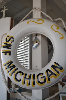 A decorative life preserver ring with 'SWF Michigan' written on it hangs from a rope. A small metallic fish ornament is attached to the center, and a window with blinds is visible in the background.