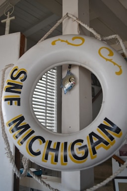 A decorative life preserver ring with 'SWF Michigan' written on it hangs from a rope. A small metallic fish ornament is attached to the center, and a window with blinds is visible in the background.