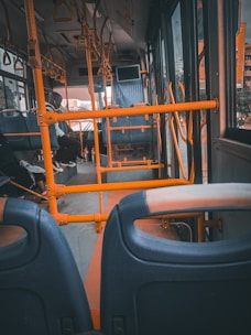 The interior of a bus featuring orange railings and blue seats, with some passengers seated and visible through the window. The bus ceiling has handles for standing passengers, and there is a screen at the front.