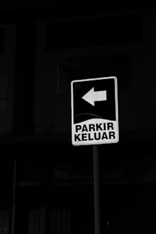 A parking exit sign is displayed, featuring a bold white arrow pointing to the left and the words 'PARKIR KELUAR' in capital letters. The background is predominantly dark, highlighting the sign.