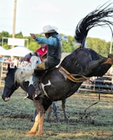 A rodeo cowboy is riding a bucking horse in an outdoor arena. The cowboy wears a white hat, blue shirt, and protective vest, while holding onto the saddle. The horse, black and white in color, is mid-buck with dirt flying around. A crowd and other riders are visible in the background.