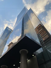 A modern glass skyscraper reflecting a partly cloudy blue sky and adjacent buildings. The structure is sleek and imposing, with a geometric design emphasizing vertical lines and transparency.