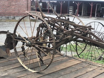 A rusted antique farming implement with large spoked wheels and metal components is placed on a wooden platform. The background features a brick building and gravel ground.