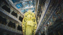 A large, ornate statue of a deity stands prominently within a richly decorated temple interior. The architecture showcases intricate details, vibrant patterns, and a high ceiling with a sky-like design. The statue is surrounded by an elaborate backdrop, emphasizing its spiritual significance.