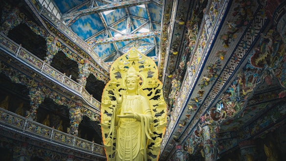 A large, ornate statue of a deity stands prominently within a richly decorated temple interior. The architecture showcases intricate details, vibrant patterns, and a high ceiling with a sky-like design. The statue is surrounded by an elaborate backdrop, emphasizing its spiritual significance.