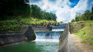 A multi-level concrete waterway with cascading water flowing down steps, surrounded by lush green vegetation and trees under a partly cloudy sky. The water is clear and the setting appears serene and natural.