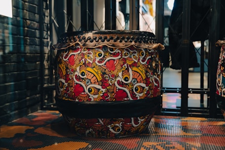 A highly decorative drum featuring a vibrant and intricate design filled with bold, colorful patterns. The design includes swirling shapes and stylized motifs in red, yellow, black, and white. The drum is situated in an industrial-looking space with metal and brick elements surrounding it.