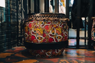 A highly decorative drum featuring a vibrant and intricate design filled with bold, colorful patterns. The design includes swirling shapes and stylized motifs in red, yellow, black, and white. The drum is situated in an industrial-looking space with metal and brick elements surrounding it.