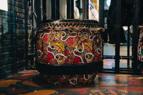 A highly decorative drum featuring a vibrant and intricate design filled with bold, colorful patterns. The design includes swirling shapes and stylized motifs in red, yellow, black, and white. The drum is situated in an industrial-looking space with metal and brick elements surrounding it.