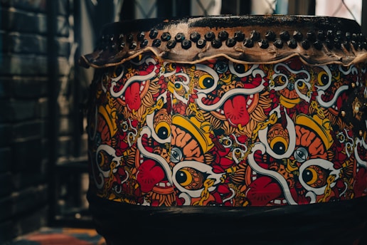 A large drum decorated with vibrant, abstract artwork featuring bold eyes, tongues, and colorful swirls. The design includes vivid patterns in red, yellow, white, and black. The top of the drum is secured with a series of black studs or rivets.