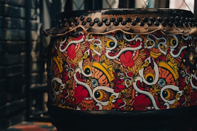 A large drum decorated with vibrant, abstract artwork featuring bold eyes, tongues, and colorful swirls. The design includes vivid patterns in red, yellow, white, and black. The top of the drum is secured with a series of black studs or rivets.