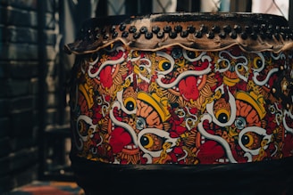A large drum decorated with vibrant, abstract artwork featuring bold eyes, tongues, and colorful swirls. The design includes vivid patterns in red, yellow, white, and black. The top of the drum is secured with a series of black studs or rivets.