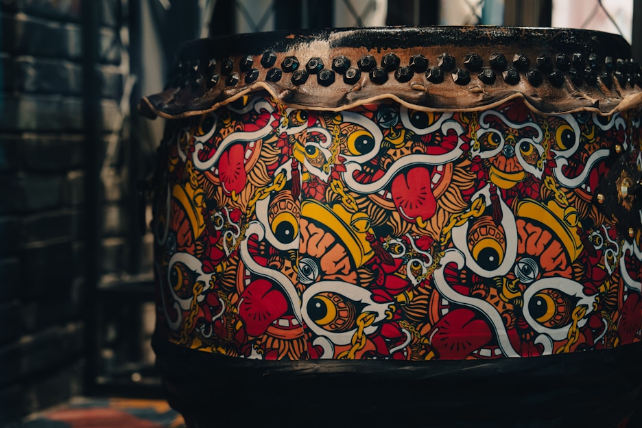 A large drum decorated with vibrant, abstract artwork featuring bold eyes, tongues, and colorful swirls. The design includes vivid patterns in red, yellow, white, and black. The top of the drum is secured with a series of black studs or rivets.