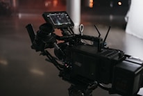 A professional video camera setup with multiple components including a monitor and various controls. The equipment features a detailed array of buttons and dials, and the environment has a subdued, low-light atmosphere with reflective surfaces.