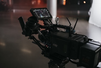 A professional video camera setup with multiple components including a monitor and various controls. The equipment features a detailed array of buttons and dials, and the environment has a subdued, low-light atmosphere with reflective surfaces.