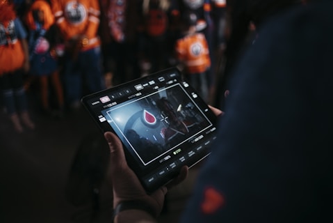 A person is holding a tablet that displays a video or image of a red and blue emblem on the screen. In the background, several people wearing dark and orange colored clothing, possibly sports jerseys, are partially visible. The scene is dimly lit, suggesting an indoor or evening setting.