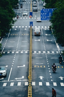 A wide, multi-lane road with a central barrier and directional arrows painted on the asphalt. There are multiple vehicles, including cars, vans, and motorcycles, traveling in various directions. The road is lined with trees and features traffic signs and signals, with some signs displayed prominently above the lanes.