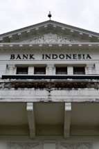 An ornate building facade displaying the words 'Bank Indonesia' in bold letters. The architecture features intricate carvings and decorative details above the entrance, with a prominent triangular pediment and Corinthian columns. The color scheme is primarily white and light gray with some weathering and black detailing.