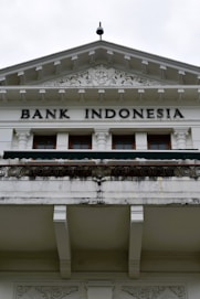 An ornate building facade displaying the words 'Bank Indonesia' in bold letters. The architecture features intricate carvings and decorative details above the entrance, with a prominent triangular pediment and Corinthian columns. The color scheme is primarily white and light gray with some weathering and black detailing.