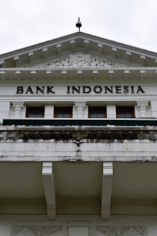 An ornate building facade displaying the words 'Bank Indonesia' in bold letters. The architecture features intricate carvings and decorative details above the entrance, with a prominent triangular pediment and Corinthian columns. The color scheme is primarily white and light gray with some weathering and black detailing.