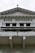 An ornate building facade displaying the words 'Bank Indonesia' in bold letters. The architecture features intricate carvings and decorative details above the entrance, with a prominent triangular pediment and Corinthian columns. The color scheme is primarily white and light gray with some weathering and black detailing.