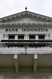 An ornate building facade displaying the words 'Bank Indonesia' in bold letters. The architecture features intricate carvings and decorative details above the entrance, with a prominent triangular pediment and Corinthian columns. The color scheme is primarily white and light gray with some weathering and black detailing.