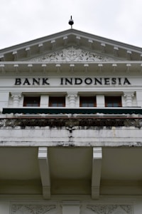 An ornate building facade displaying the words 'Bank Indonesia' in bold letters. The architecture features intricate carvings and decorative details above the entrance, with a prominent triangular pediment and Corinthian columns. The color scheme is primarily white and light gray with some weathering and black detailing.