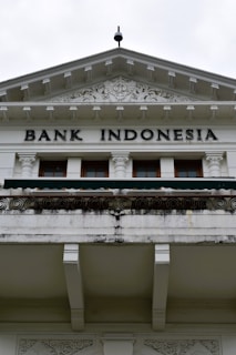 An ornate building facade displaying the words 'Bank Indonesia' in bold letters. The architecture features intricate carvings and decorative details above the entrance, with a prominent triangular pediment and Corinthian columns. The color scheme is primarily white and light gray with some weathering and black detailing.