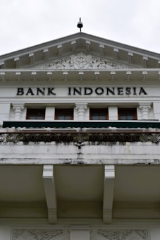 An ornate building facade displaying the words 'Bank Indonesia' in bold letters. The architecture features intricate carvings and decorative details above the entrance, with a prominent triangular pediment and Corinthian columns. The color scheme is primarily white and light gray with some weathering and black detailing.