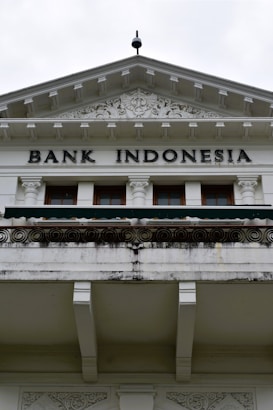 An ornate building facade displaying the words 'Bank Indonesia' in bold letters. The architecture features intricate carvings and decorative details above the entrance, with a prominent triangular pediment and Corinthian columns. The color scheme is primarily white and light gray with some weathering and black detailing.