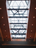 A view is directed upwards at a modern ceiling with large geometric glass panels. The ceiling features a grid-like pattern and a large ceiling fan in the center. The surrounding edges are illuminated with small recessed lights, creating a warm and inviting atmosphere.