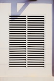 A rectangular vent is mounted on a wall, featuring evenly spaced horizontal slats. The surrounding area is a light-colored surface with a shadow cast across it.