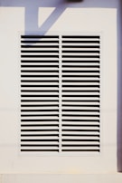 A rectangular vent is mounted on a wall, featuring evenly spaced horizontal slats. The surrounding area is a light-colored surface with a shadow cast across it.