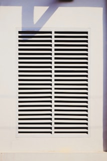 A rectangular vent is mounted on a wall, featuring evenly spaced horizontal slats. The surrounding area is a light-colored surface with a shadow cast across it.