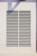 A rectangular vent is mounted on a wall, featuring evenly spaced horizontal slats. The surrounding area is a light-colored surface with a shadow cast across it.