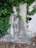 A stone relief sculpture depicts a solemn-looking person holding a smaller figure. The artwork is surrounded by lush green ivy climbing up a weathered stone wall. The setting suggests a serene and slightly mysterious atmosphere.