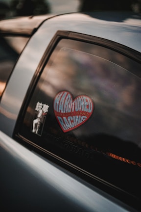 A close-up view of a car window displaying a heart-shaped sticker with the words 'HARDTUNED RACING' and another smaller sticker with text and an illustration.