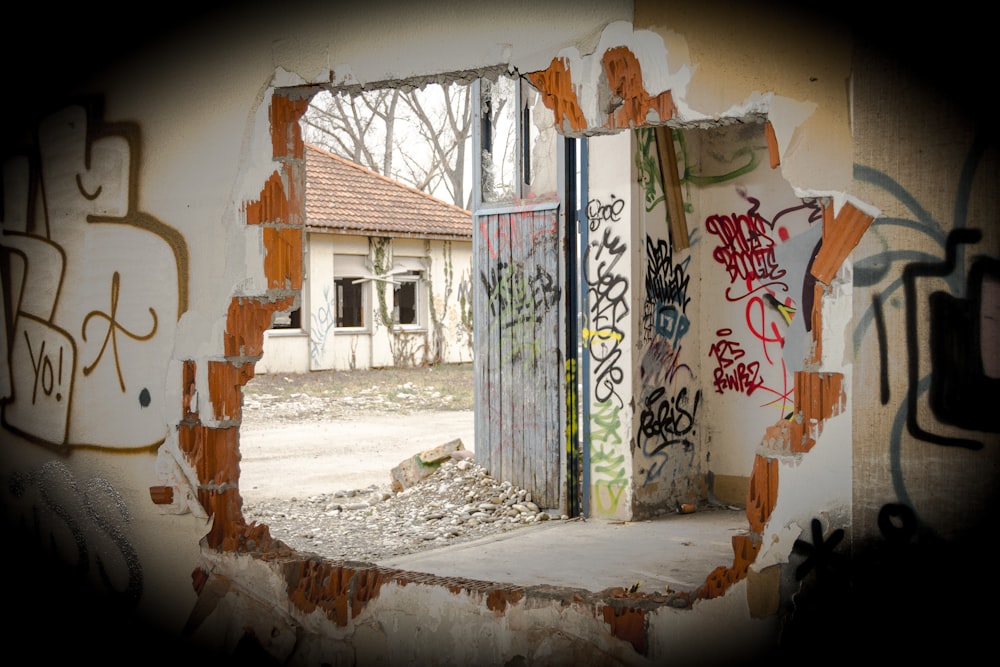 a door with graffiti on it is seen through a hole in a wall